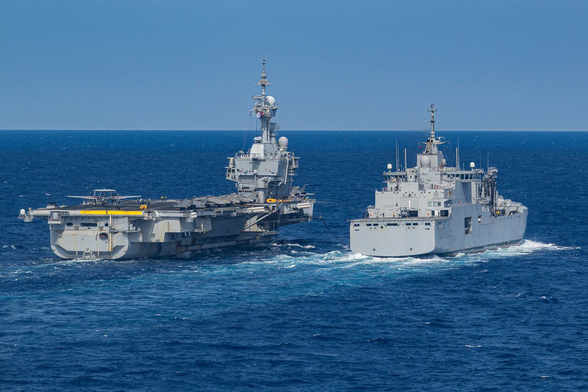 Replenishment-at-sea constrains the carrier to a slower speed range of about 12 to 16 knots and a fixed heading with minimal deviation, preventing it from turning into the wind or accelerating to generate an optimal airflow for the E-2C. (Picture source: French Navy)