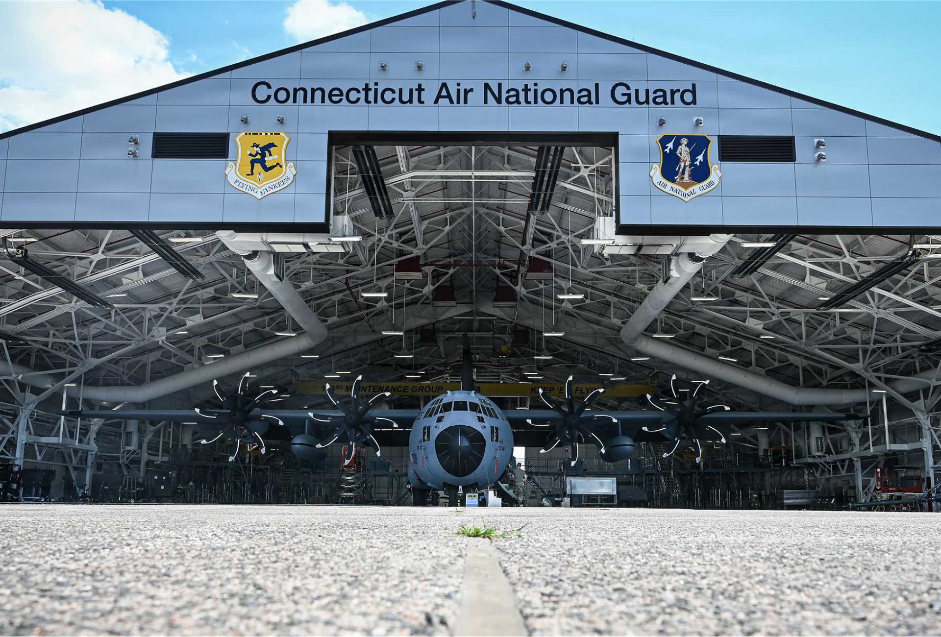 Airmen from the 103rd Maintenance Group, U.S. Air National Guard, open the hangar doors on a clear day to unveil a fully upgraded C-130H Hercules military transport aircraft equipped with NP2000 propellers and T56 3.5 engines.