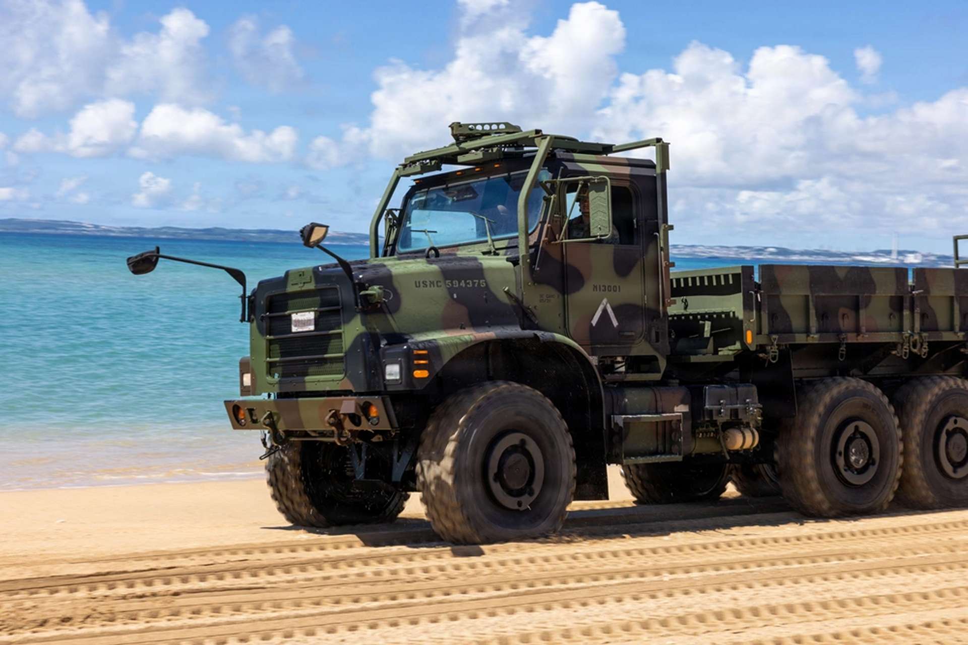 U.S. Marines operate MTVR 6x6 tactical trucks during littoral mobility training at Kin Blue Beach, Okinawa, testing movement across soft sand, unimproved routes and vehicle self-recovery in support of dispersed expeditionary operations (Picture source: U.S. DoW).