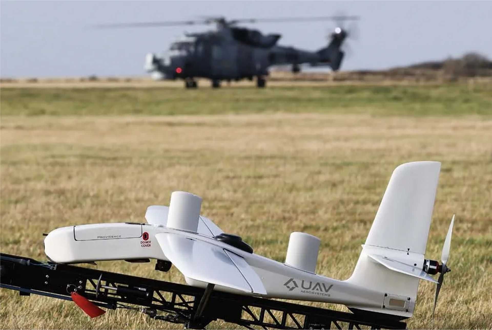 A British Royal Navy unmanned aerial system is positioned on the ground as an AW159 Wildcat helicopter prepares for flight in the background during the Eagles Eye trials, highlighting crewed-uncrewed integration and network-enabled operations.