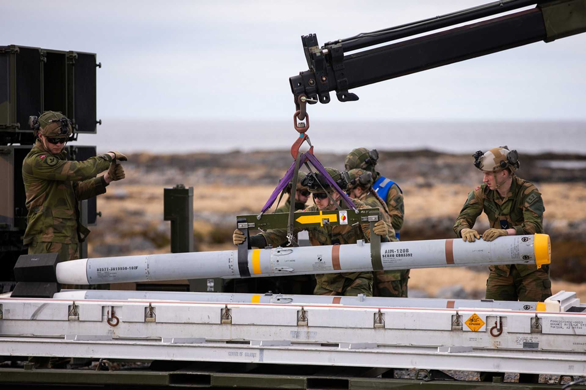 An AIM-120 AMRAAM missile is loaded onto a NASAMS launcher unit during a NATO air defense readiness exercise. The system is designed for rapid engagement of aerial threats including cruise missiles, drones, and aircraft using networked radar and precision-guided interceptors.