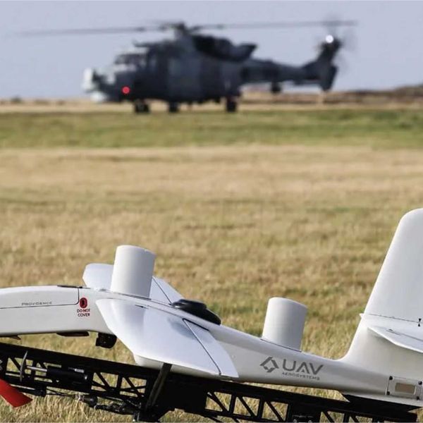 A British Royal Navy unmanned aerial system is positioned on the ground as an AW159 Wildcat helicopter prepares for flight in the background during the Eagles Eye trials, highlighting crewed-uncrewed integration and network-enabled operations.